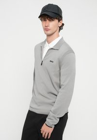 Młody mężczyzna w czarnej czapce, szarym swetrze z półzamek i logo "BOSS", białej koszuli z kołnierzykiem pod spodem oraz czarnych spodniach na jednolitym tle.