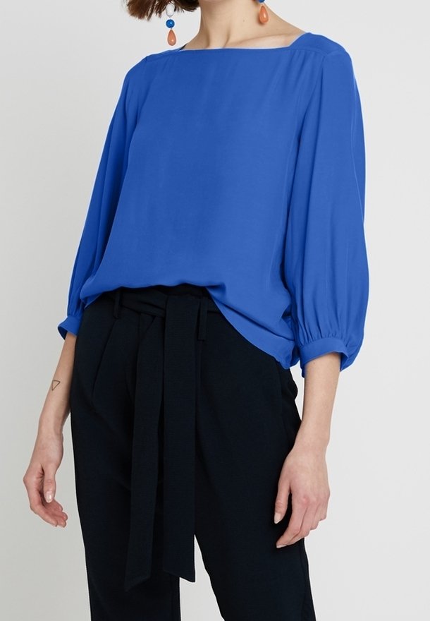 Blouse bleu ample avec un décolleté carré, des manches trois-quarts froncées et une texture lisse, assortie à un pantalon noir taille haute.