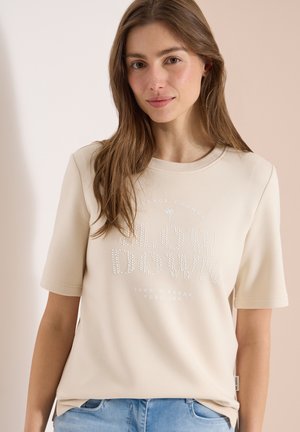 Jeune femme aux longs cheveux châtain clair portant un T-shirt beige avec le texte "SLOW DOWN" et un jean bleu clair, debout devant un fond neutre.