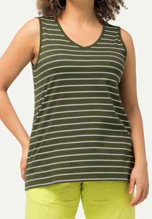 Frau trägt ärmelloses olivgrünes und graues gestreiftes Oberteil mit V-Ausschnitt und leuchtend gelbe Hose, steht mit einer Hand in der Hüfte.