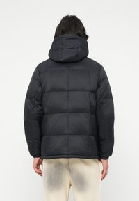 Μαύρο μπουφάν τύπου puffer με �κουκούλα, καπιτονέ σχέδιο και ελαστικές μανσέτες. Το υλικό φαίνεται ελαφρύ και μονωμένο για ζεστασιά.