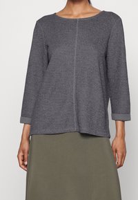 Graues Langarmshirt mit gerippter Textur, weitem Rundhalsausschnitt und hochgerollten Ärmeln. Gerader Schnitt mit einer Mittelnaht-Detail.