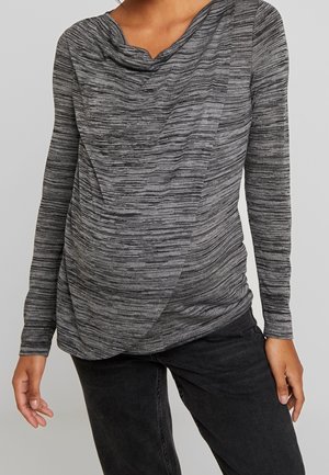 Donna che indossa un top grigio a maniche lunghe, testurizzato, con scollo drappeggiato e jeans neri, in piedi con le braccia rilassate ai lati contro uno sfondo neutro.