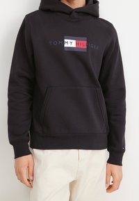 Black hooded sweatshirt with a front kangaroo pocket, featuring a white and red rectangular logo on the chest. Smooth fabric texture.