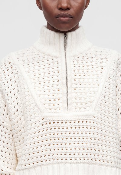 Personne portant un pull blanc en tricot avec un col haut côtelé et une fermeture éclair argentée partiellement fermée à l'avant.