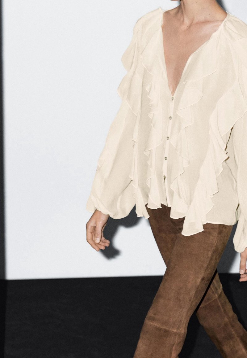 Femme portant un chemisier crème à volants avec des boutons et un pantalon en daim marron, vue du cou jusqu’aux cuisses sur un fond uni.