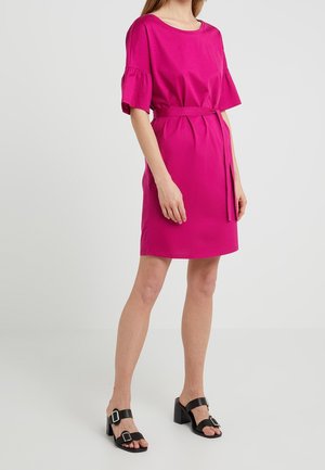 Kobieta w jaskrawo różowej sukience do kolan z wiązaniem w talii i krótkimi, falbankowymi rękawami, w zestawieniu z czarnymi sandałami na obcasie z klamrami.