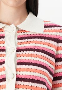 Artimiausiu planu matoma asmuo, kuris dėvi dryžuotą megztinį rožinės, oranžinės ir grietinėlės spalvų, su apykakle ir mygtukų detalėmis.