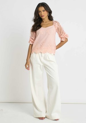 Woman with long dark hair wears a sheer pink embroidered blouse with puff sleeves and white wide-leg trousers, standing with one hand on hip.
