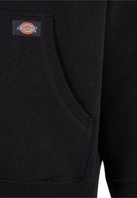 Black sweatshirt featuring a front pocket, soft cotton material, and a woven label with a circular logo. Ribbed accents at hem and cuffs.