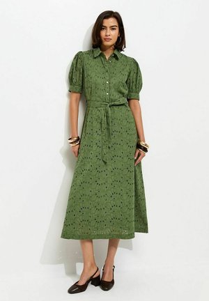 Kobieta w zielonej sukience midi z guzikami, krótkimi bufkami, paskiem w talii, czarnych butach na obcasie typu slingback oraz złotych bransoletkach na obu nadgarstkach.
