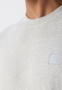 Jasnoszara bluza z ribbowanym dekoltem, z małym wytłoczonym logo w kwadratowej naszywce na klatce piersiowej. Miękka tekstura, casualowy design.