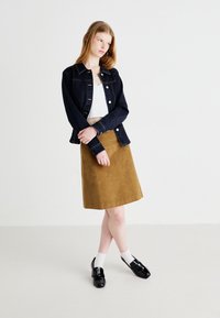 Veste en denim, bleu foncé, avec des boutons et des poches. Jupe en velours côtelé marron, texture lisse. Chaussures noires, chaussettes blanches, debout sur un fond clair.