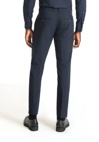 Pantaloni da abito blu realizzati in tessuto liscio, con una vestibilità su misura, due tasche posteriori con bottone e un design a gamba affusolata. Scarpe nere mostrate.