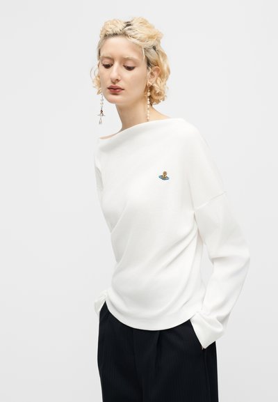 Γυναίκα με κοντά κατσαρά ξανθά μαλλιά φοράει λευκό μακρυμάνικο τοπ με μικρό πολύχρωμο έμβλημα στο στήθος και μαύρο παντελόνι, με τα χέρια στις τσέπες.