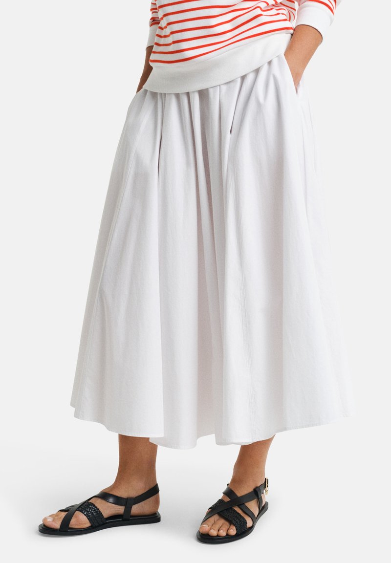 Jupe midi plissée blanche avec une texture lisse, associée à des sandales noires à lanières. Le haut présente des rayures horizontales rouges et blanches.