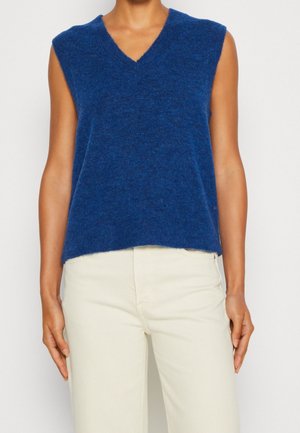 Personne portant un gilet sans manches bleu à col en V et un pantalon taille haute beige clair sur un fond uni.