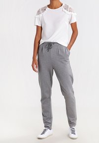 Haut à manches courtes blanc avec des accents en dentelle sur les épaules, associé à un pantalon de jogging gris à cordon et des baskets blanches. Tenue décontractée et confortable.