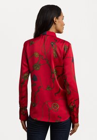Red silk blouse featuring a printed design of gold ropes and emblems, with a buttoned collar and long sleeves. Smooth texture, relaxed fit.