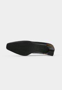 Chaussure plate noire à bout pointu posée sur le côté, montrant une semelle lisse et un petit talon fin avec un accent doré à l'arrière.