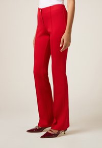 Rode op maat gemaakte broeken met een uitlopende silhouette, voorzien van een ritssluiting en contrasterende stiksels. Gecombineerd met puntige bordeauxrode hakken.