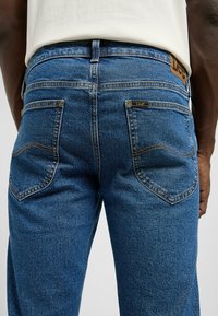 Pantalones vaqueros de denim azul con corte recto, que presentan costuras amarillas, una etiqueta de cuero en la cintura y dos bolsillos traseros con costuras curvadas.