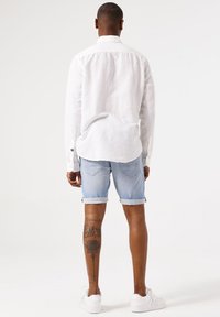 Witte linnen blouse met lange mouwen, lichtblauwe denim shorts, witte sneakers, zichtbare bloemen tattoo op het been, opgerolde shorts, relaxed fit.