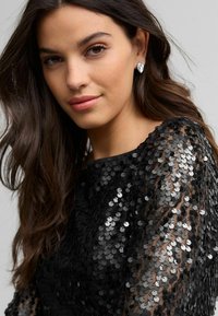 Top in paillettes nere con manica trasparente, caratterizzato da paillettes tonde per texture. La modella sfoggia un trucco neutro e un sottile dettaglio di orecchini.