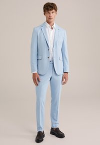 Ljusblå kostym bestående av en skräddarsydd kavaj och slim-fit byxor, kombinerad med en vit skjorta med knappar och mörkbruna loafers.