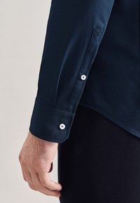 Camicia a maniche lunghe blu navy con polsino abbottonato, caratterizzata da una texture liscia, bottoni bianchi e un ricamo sottile vicino al polsino.