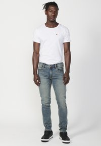 Fehér, kerek nyakú póló, világoskék, megviselt farmernadrág és fekete sneaker. Egyszerű dizájn, pamut anyag, slim fit szabás és minimális márkázás.