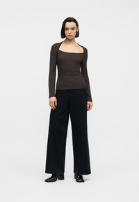 Γυναίκα με κοντά σκουρόχρωμα μαλλιά, φορώντας εφαρμοστό σκούρο καφέ τοπ με μακριά μανίκια και τετράγωνη λαιμόκοψη, φαρδιά μαύρα παντελόνια και μαύρα μποτάκια με τακούνια.