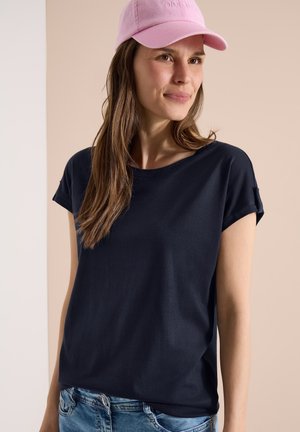 Femme aux longs cheveux bruns portant une casquette de baseball rose, un t-shirt bleu marine à manches courtes et un jean bleu sur un fond beige.