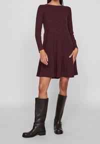 Žebrované vínové šaty s dlouhými rukávy a rozšířenou sukní. Kombinováno s černými koženými kozačkami po kolena s detailním sponou.