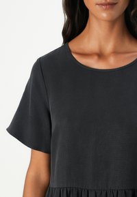 Schwarzes Kurzarmkleid mit rundem Ausschnitt und texturiertem Stoff, das am Saum gerafft ist, um zusätzliche Details zu bieten.