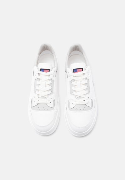 Paire de baskets blanches avec lacets et un petit drapeau américain sur la languette, vues de dessus sur un fond blanc.