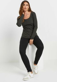 Tumma oliivinvihreä pitkähihainen ribbipaita, jossa on pyöreä pääntie ja nappi etupaneelissa, yhdistettynä mustiin legginsseihin ja valkoisiin tennareihin.