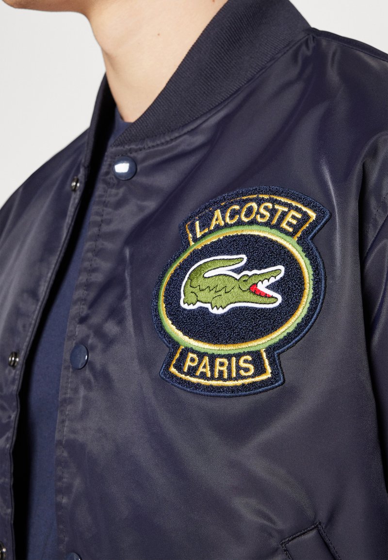 Veste bomber en satin bleu marine avec un logo crocodile Lacoste brodé en vert et blanc, un patch rond avec le texte "PARIS" et des fermetures à pression.