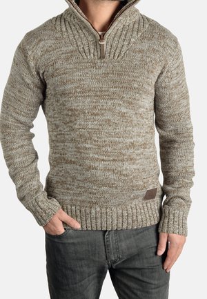 Man wearing a beige and brown speckled knit sweater with a high collar and half zipper, paired with dark gray jeans, hands relaxed.