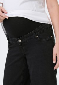 Gravid person iført sorte graviditetsjeans med elastik i taljen under en hvid skjorte, løfter skjorten for at vise mavepanelet.