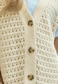 Gilet en tricot de couleur crème avec un tissage ouvert texturé, présentant quatre grands boutons marron sur le devant et un design en V.