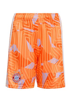 Ung dreng, der står med hænderne bag ryggen iført en skarp orange fodbolduniform med sorte knopper på ensfarvet baggrund.