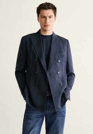 Mann trägt einen marineblauen zweireihigen Blazer, ein dunkelblaues Hemd und blaue Jeans und steht vor einem schlichten hellen Hintergrund.