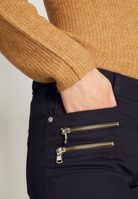 Mustat housut, joissa kultaiset vetoketjulliset taskut; kangas vaikuttaa sileältä ja kevyesti kiiltävältä. Käsi on oikeassa taskussa, mikä korostaa yksityiskohtia.