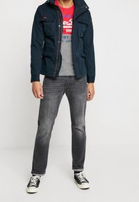 Mörkblå zip-up jacka med huva, två framfickor, bärs över en grå skjorta med röd grafik, i kombination med grå jeans och svarta sneakers.