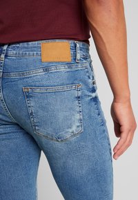 Slim fit blå jeans i denim med en blekt tvättning, med en ljusbrun läderetikett vid midjebandet och vanliga bakfickor.