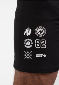 Zwarte shorts met witte geborduurde logo's, waaronder gorillagraphics, cirkelvormige emblemen, het nummer "82" en gekruiste initialen "GW."