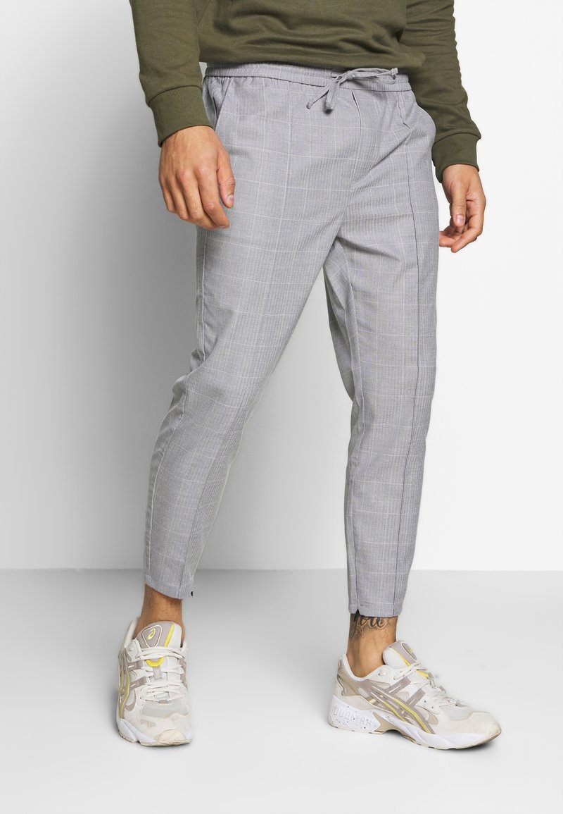 Homme portant un pantalon à carreaux gris clair avec cordon, un sweat-shirt vert olive et des chaussures de sport blanches avec des touches jaunes, debout sur un sol blanc.