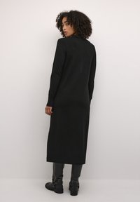 Long black knit dress with long sleeves, a straight cut, and ribbed cuffs, paired with black ankle boots featuring buckles.