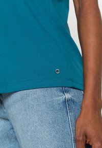 Camisa de manga corta color teal con un dobladillo curvado, que presenta un pequeño detalle de logotipo plateado cerca de la parte inferior, combinada con jeans de mezclilla azul claro.